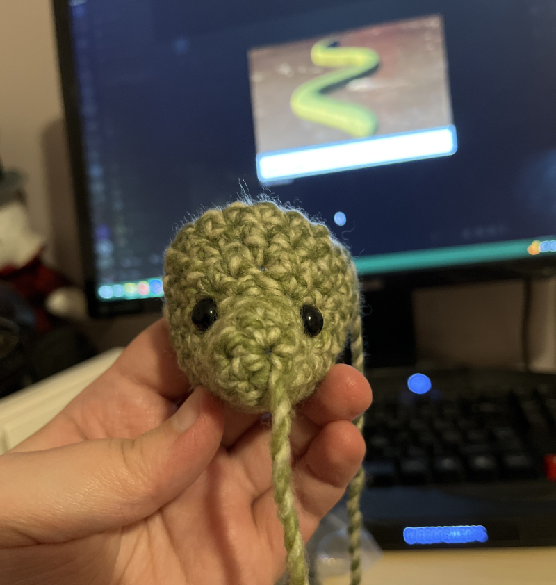 the face of a plush dinosaur with beady, black eyes. it is crocheted out of green-and-biege yarn. a blurred desktop monitor is visible in the background with an image of a cartoony green snake pulled up in the center.