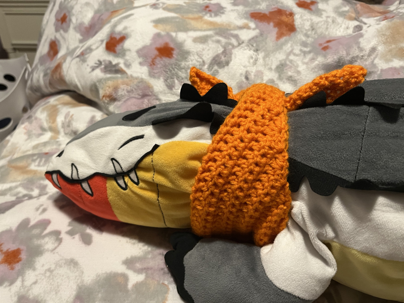 a profile view of a plush alligator wearing an orange bandana around its neck. the gator itself is white, grey, and black, with red, orange, and yellow stripes on its belly.