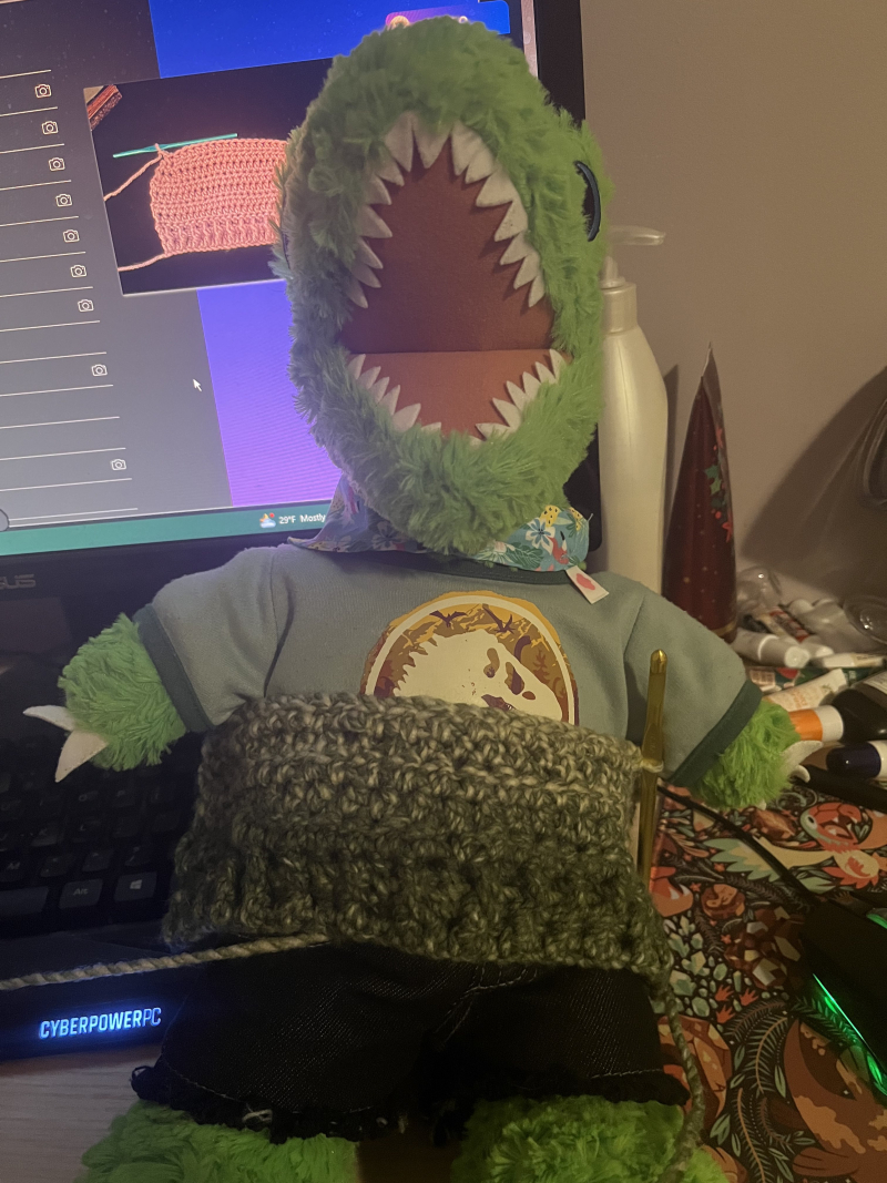 the bottom half of a miniature green sweater resting on a plush t-rex to gauge the fit. a crocheting tutorial is visible on a desktop monitor behind it.