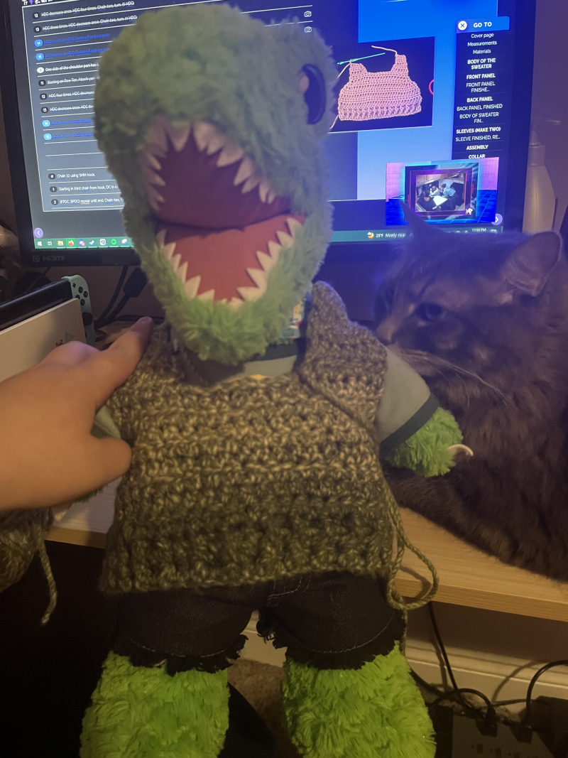 the front of a miniature green sweater resting on a plush t-rex to gauge the fit. a crocheting tutorial is visible on a desktop monitor behind it. a cat is sniffing it from the side.