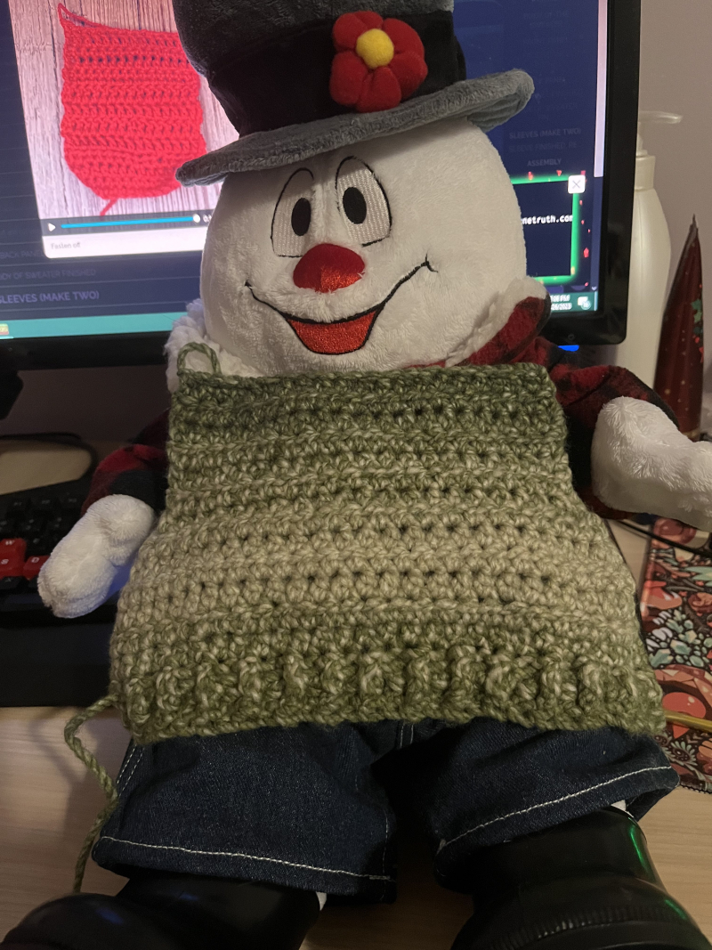 the back half of a miniature green sweater resting on a frosty the snowman plush to gauge the fit. a crocheting tutorial is visible on a desktop monitor behind it.