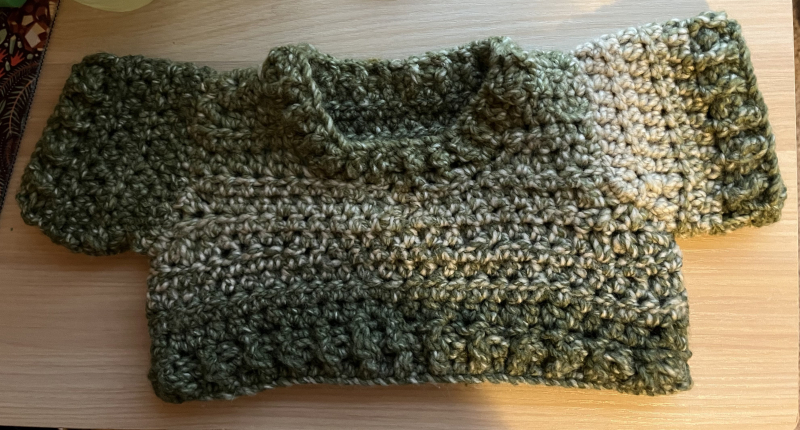 a miniature sweater crocheted out of green yarn. it is laying flat on a wooden desk.