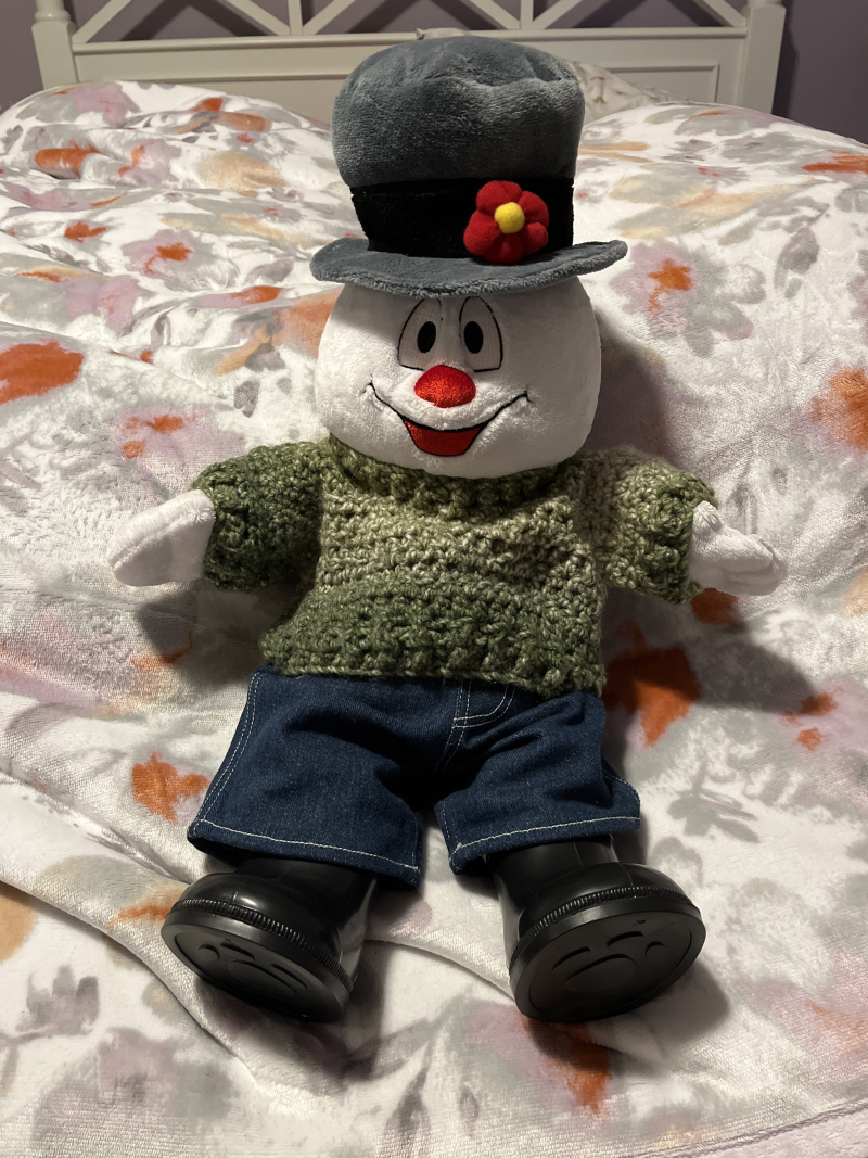a frosty the snowman plushie wearing the sweater.  he is sitting on a bed covered with a white-and-purple floral blanket.