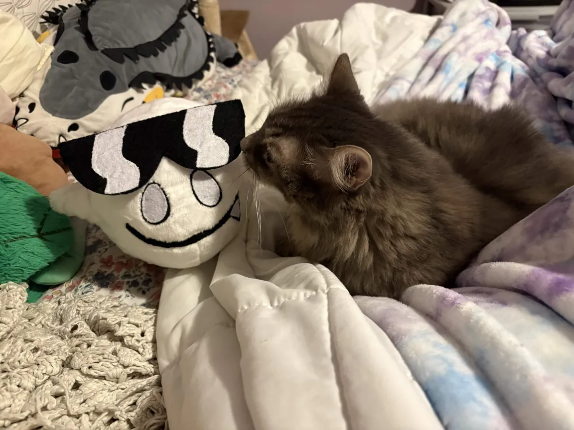 photo of a cat sniffing a plush ghost on an unmade bed. the plushie resembles a boo from the super mario series with a pair of stylized sunglasses.