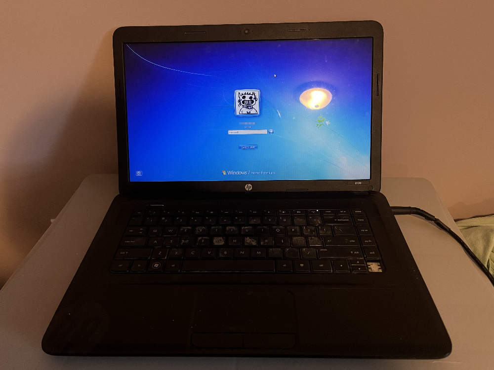 a photo of an old, black laptop on a grey surface. it has a dirty keyboard that is missing a key. the windows 8 user login screen is displayed, showing that the user's icon is a portrait of zacharie from the game OFF.