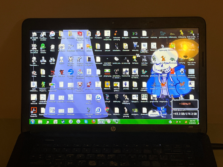 a photo of an old, black laptop on a grey surface. the desktop is visible, showing that it is overflowing with art and miscellaneous folders and program shortcuts. some of the folders use the portraits of various OFF characters for icons. the background is sans' steam trading card art, in which he is standing asleep beside a spotlight.